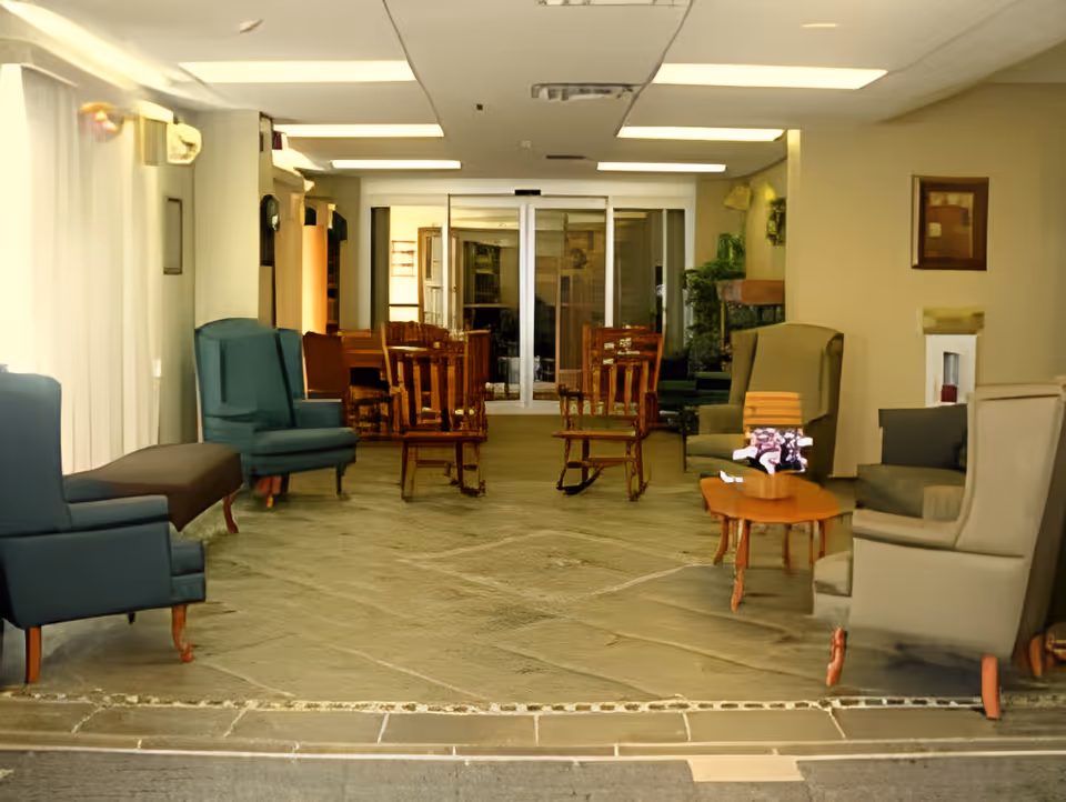 A sitting area in an assisted living facility with armchairs, wooden rocking chairs, small tables, and sliding glass doors at the far end.