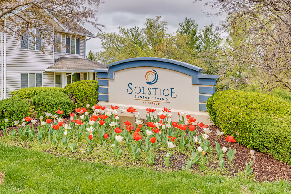 Outdoor view of the entrance sign for Solstice Senior Living at Fenton, surrounded by neatly trimmed bushes and a flower bed with red and white tulips, with part of a residential building visible in the background under a cloudy sky.