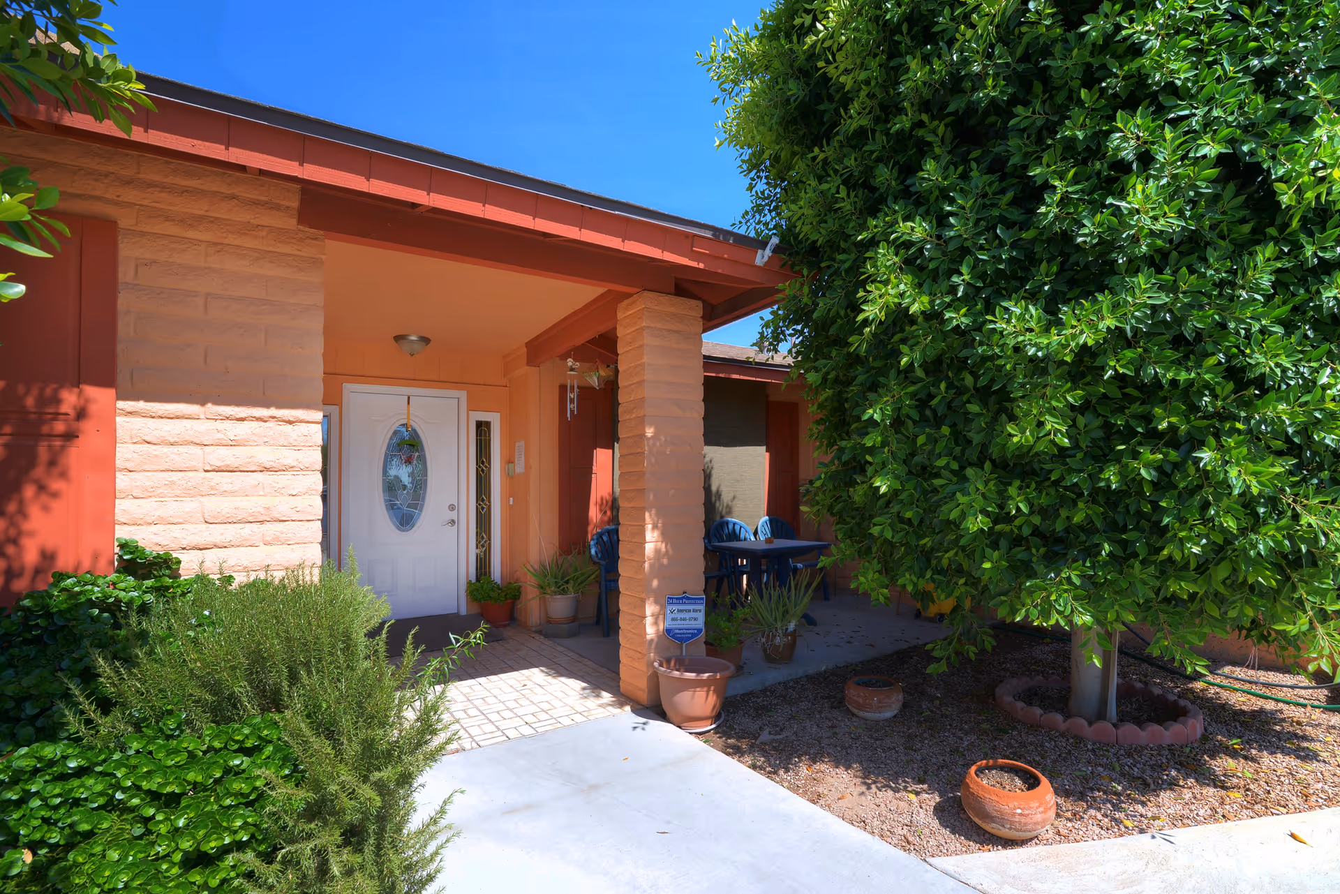 Front entrance of a single-story building with a covered porch, white door with an oval glass window, surrounded by potted plants and greenery including a large tree on the right side under a clear blue sky.