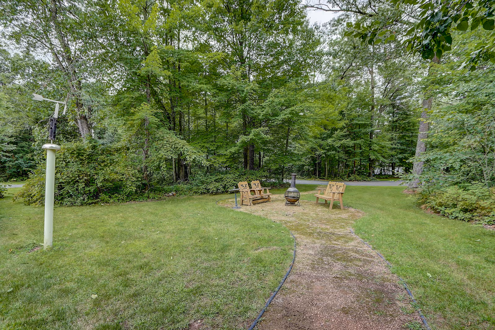 A peaceful outdoor area with a curved gravel path leading to a small seating area featuring three wooden benches arranged around a metal chiminea. The area is surrounded by lush green grass and dense trees in the background.