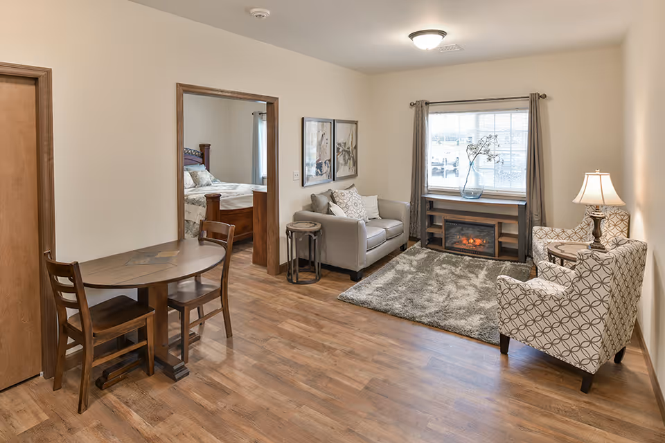 A cozy living area in a retirement community apartment featuring a small wooden dining table with two chairs, a gray loveseat with decorative pillows, two patterned armchairs, a side table with a lamp, a window with gray curtains, and a wooden console with an electric fireplace and a vase with branches. A bedroom with a wooden bed frame is visible through an open doorway.