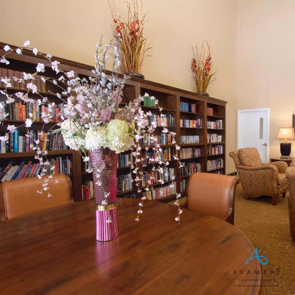 A cozy senior living common area featuring a large wooden table with a tall vase of decorative flowers in the foreground. Behind the table is a tall bookshelf filled with books and decorative vases on top. To the right, there are upholstered armchairs and a side table with a lamp, all set against beige walls.