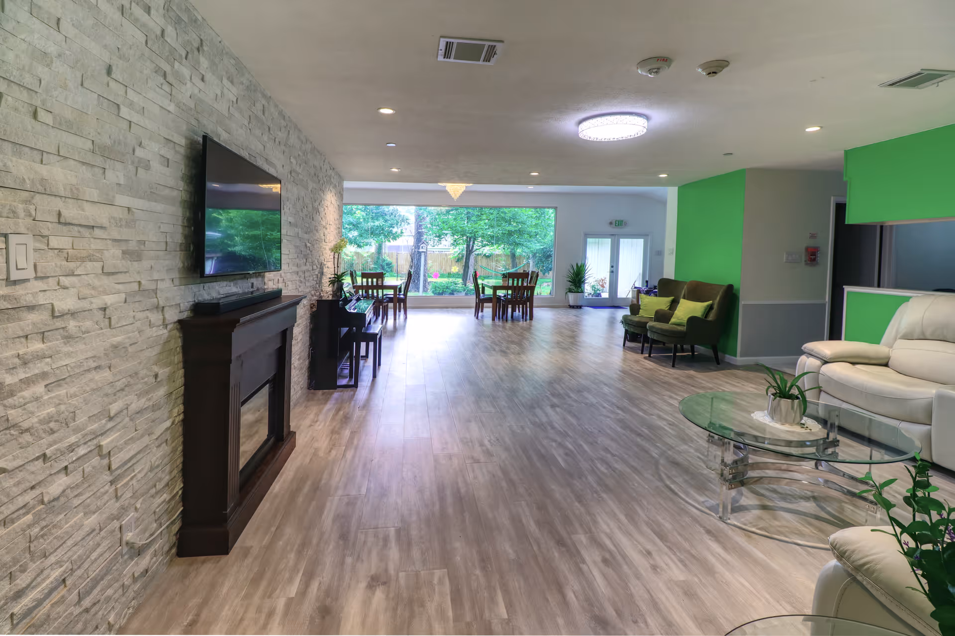 A spacious and well-lit common area in an assisted living facility featuring a modern stone accent wall with a mounted flat-screen TV and a dark wooden fireplace below it. The room has light wood flooring and a large window at the far end showing green trees outside. There are several seating areas including a green loveseat with matching pillows, a white leather sofa, and a glass coffee table with a small plant. In the background, there are dining tables and chairs near the window, and the walls have bright green accent sections.