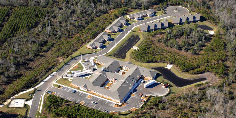 Aerial view of Grand Villa of Palm Coast senior living facility showing multiple buildings surrounded by trees and greenery, with parking lots, roads, and small ponds visible within the property.
