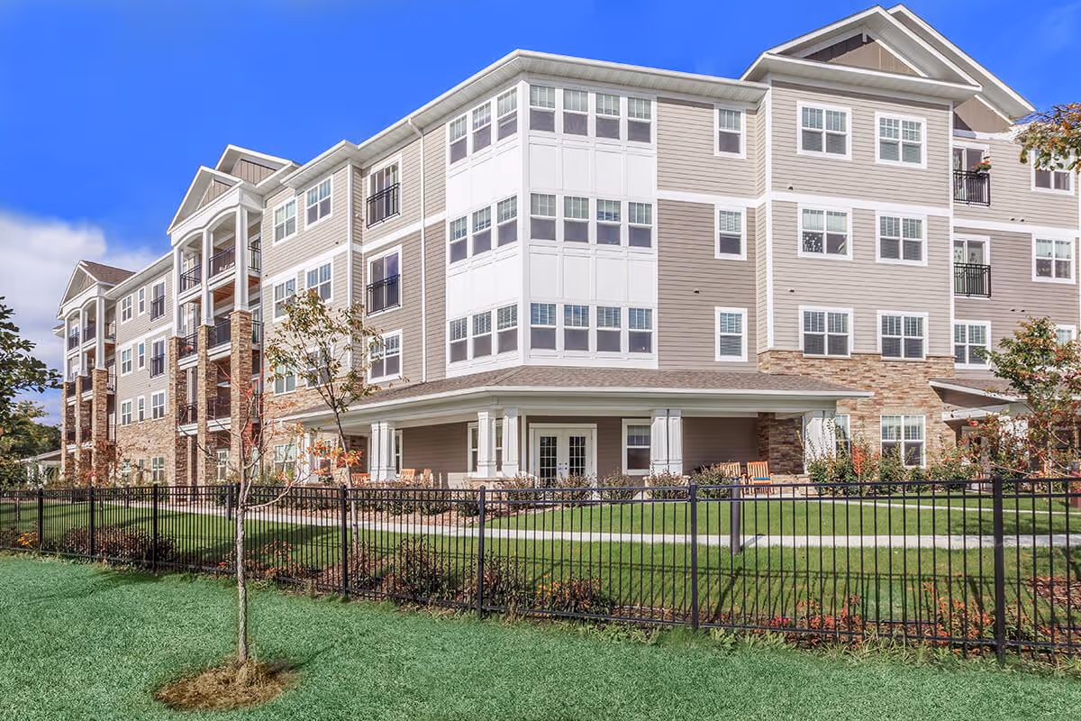 Front exterior of a four-story assisted living building with a fenced lawn and patio seating.