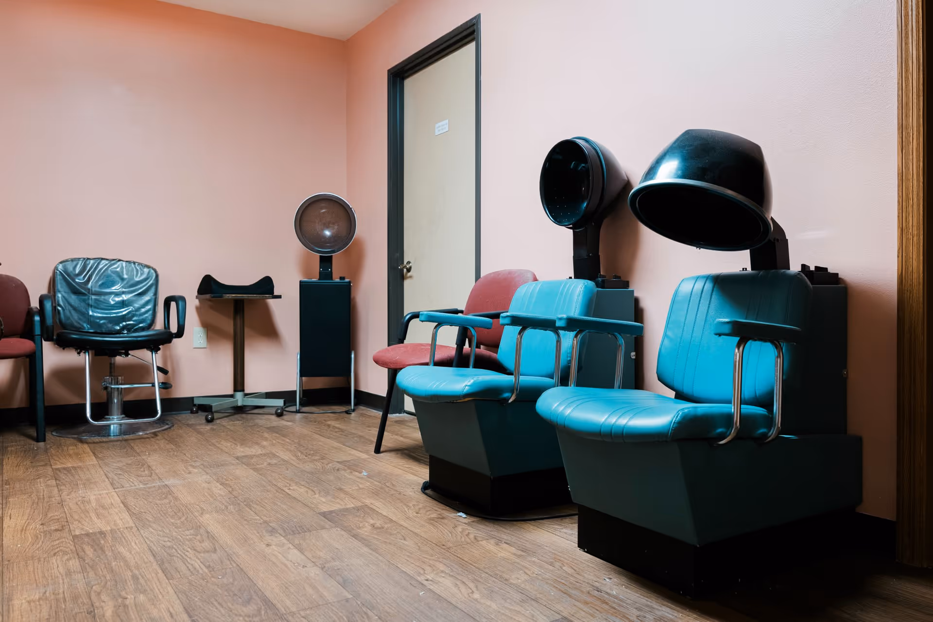 Interior of a salon area with two vintage blue hair drying chairs, a black salon chair with a plastic cover, a red chair, and a peach-colored wall with a closed door.
