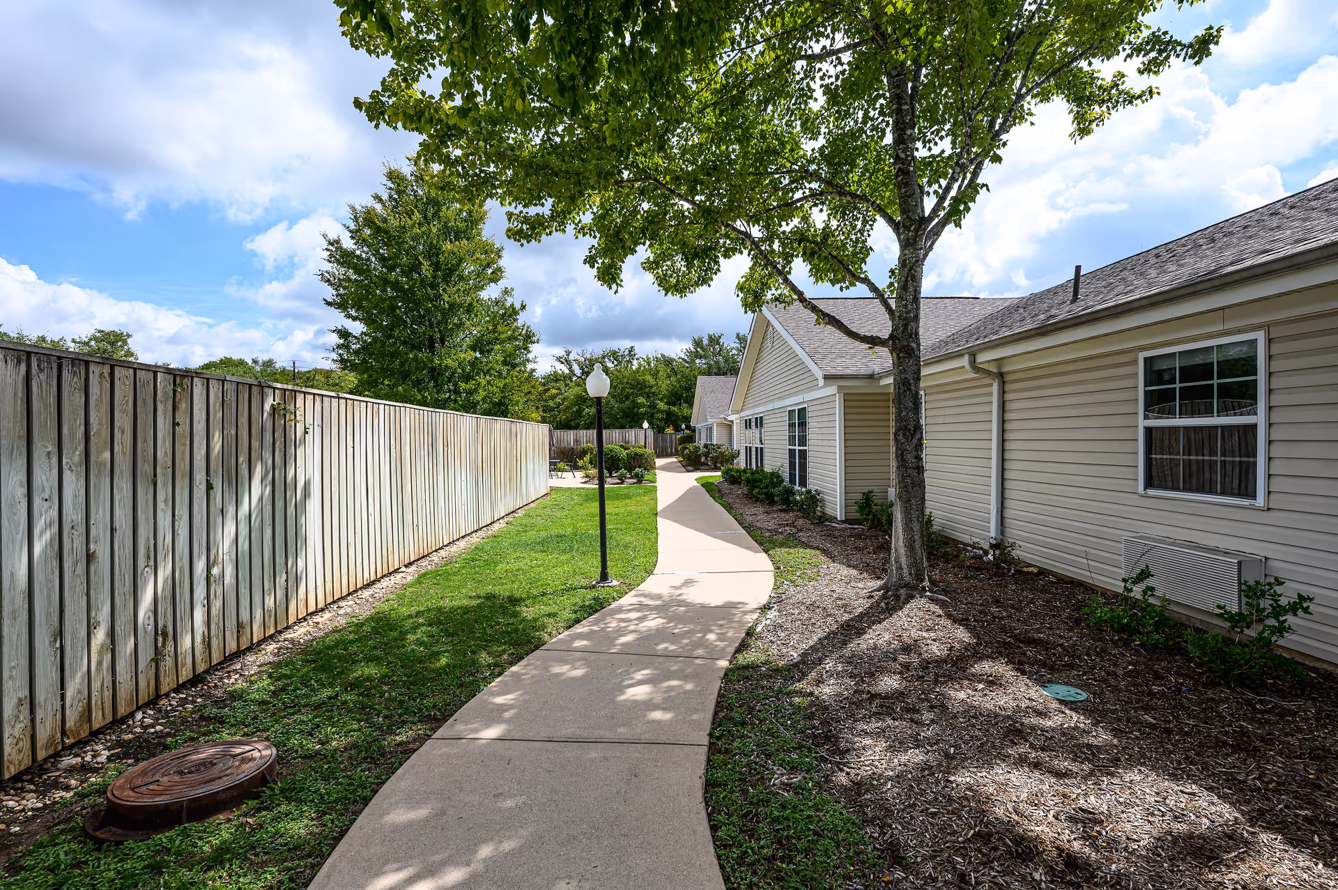A paved walkway curves through a grassy outdoor area beside a beige building with several windows. A wooden fence runs along the left side of the walkway, and a tree with green leaves provides some shade. A lamp post is positioned along the path, and the sky is partly cloudy.