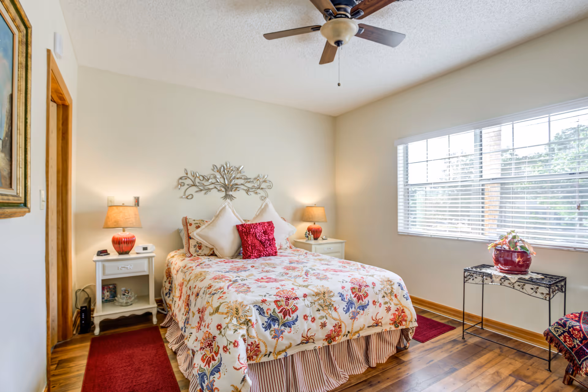 A cozy bedroom with a floral patterned bedspread on a double bed, two white nightstands with matching red lamps on either side, a decorative metal wall art above the bed, a ceiling fan, a large window with white blinds letting in natural light, a small table with a red vase and plant, and wooden flooring with a red rug.