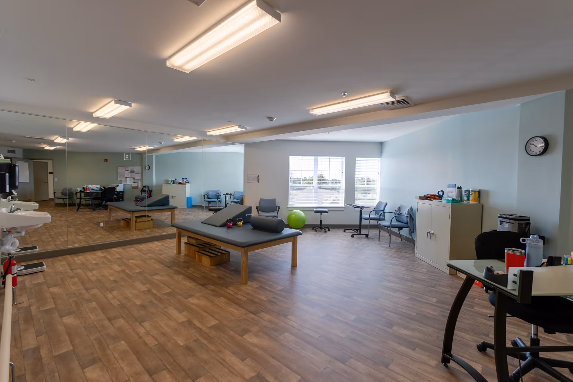 A spacious therapy or exercise room with wooden flooring, two padded therapy tables, several chairs, a large wall mirror, a green exercise ball, and a desk with office supplies. The room is well-lit with ceiling lights and natural light from a window.