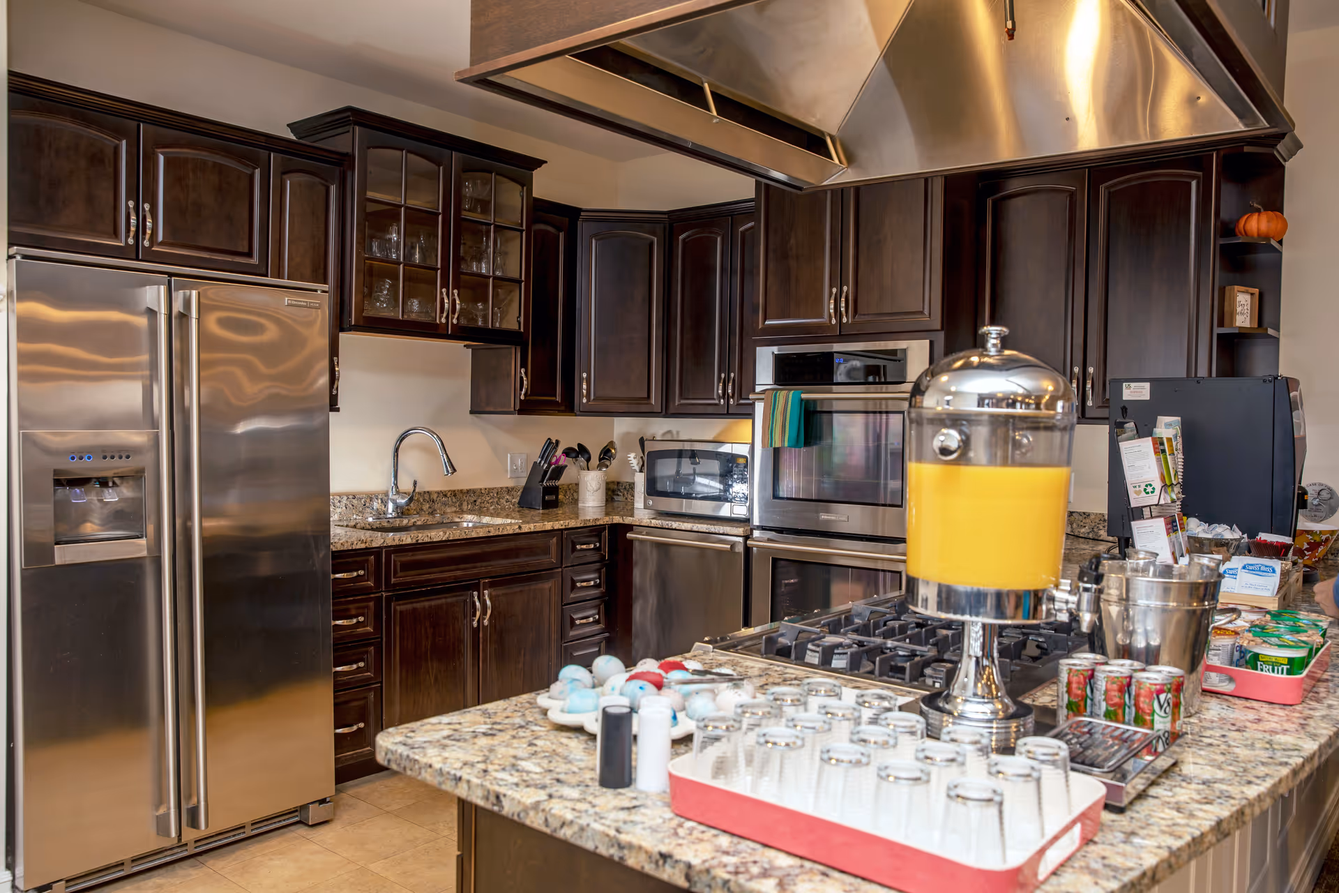 Modern communal kitchen with dark wood cabinets, stainless steel appliances, granite countertops and an island holding a large beverage dispenser and glasses.