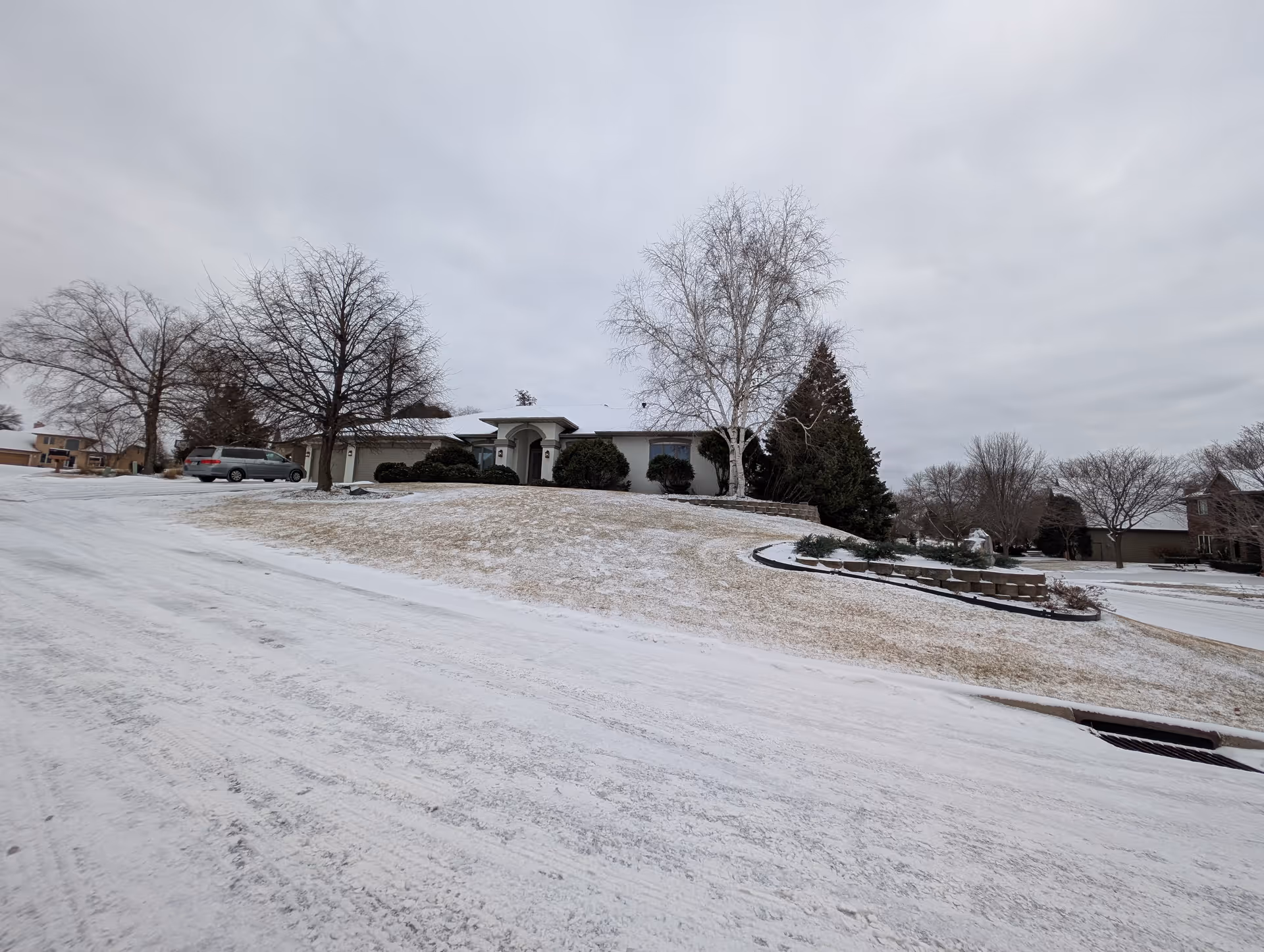 A single-story residential building on a snowy day with a sloped lawn partially covered in snow. Leafless trees and shrubs surround the house, and a gray vehicle is parked in the driveway. The sky is overcast with gray clouds.
