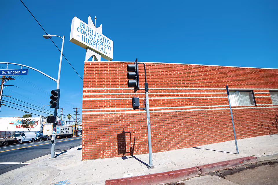 Exterior view of Burlington Convalescent Hospital showing a red brick building with a large sign on top displaying the hospital name. A traffic light and street sign for Burlington Avenue are visible at the corner, with a clear blue sky in the background.