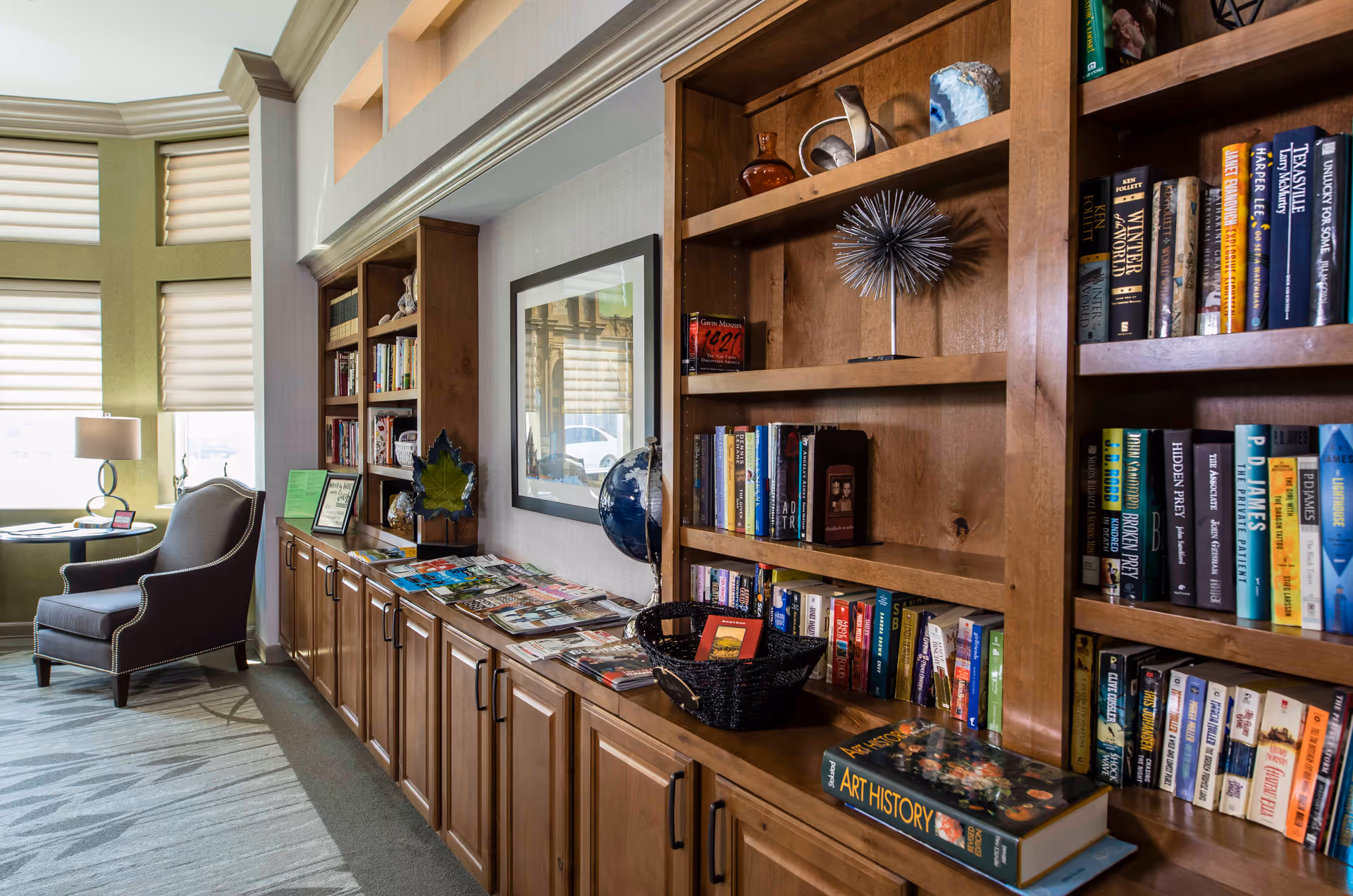 A cozy reading nook in a senior living facility featuring wooden bookshelves filled with books and decorative items, a comfortable armchair next to a round table with a lamp, and large windows with white blinds allowing natural light to fill the space.