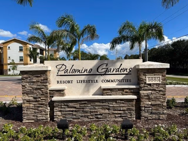 Stone and concrete sign for Palomino Gardens Resort Lifestyle Communities with palm trees and a residential building in the background under a blue sky with clouds.