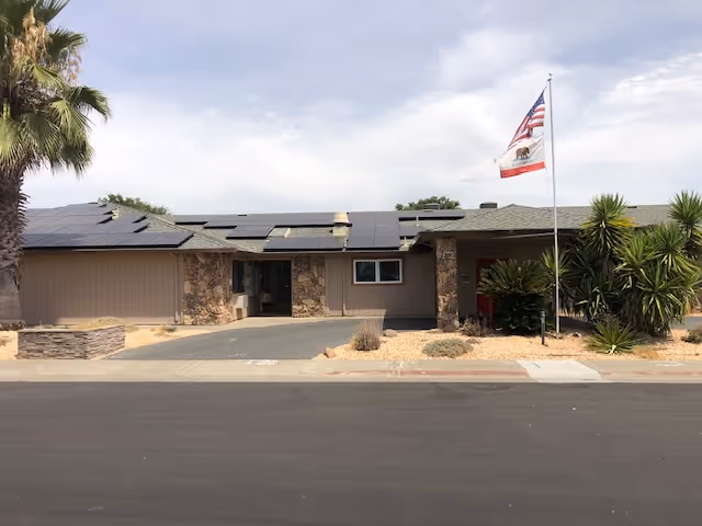 Single-story building with stone and wood exterior, solar panels on the roof, an American flag and California state flag on a flagpole in front, surrounded by desert landscaping with palm trees and shrubs under a partly cloudy sky.