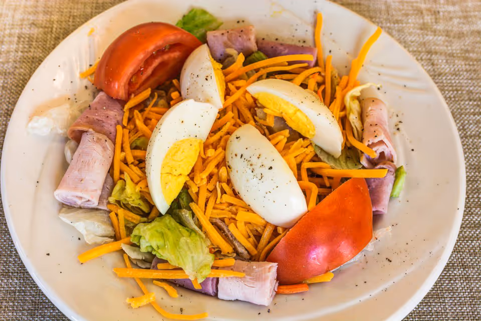Plate of salad with shredded cheddar, quartered hard-boiled eggs, tomato wedges, rolled ham, and lettuce.