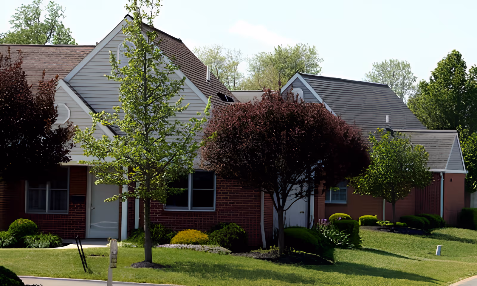 Exterior view of a residential building with brick walls and gray roofs, surrounded by green grass, trees, and shrubs on a sunny day.