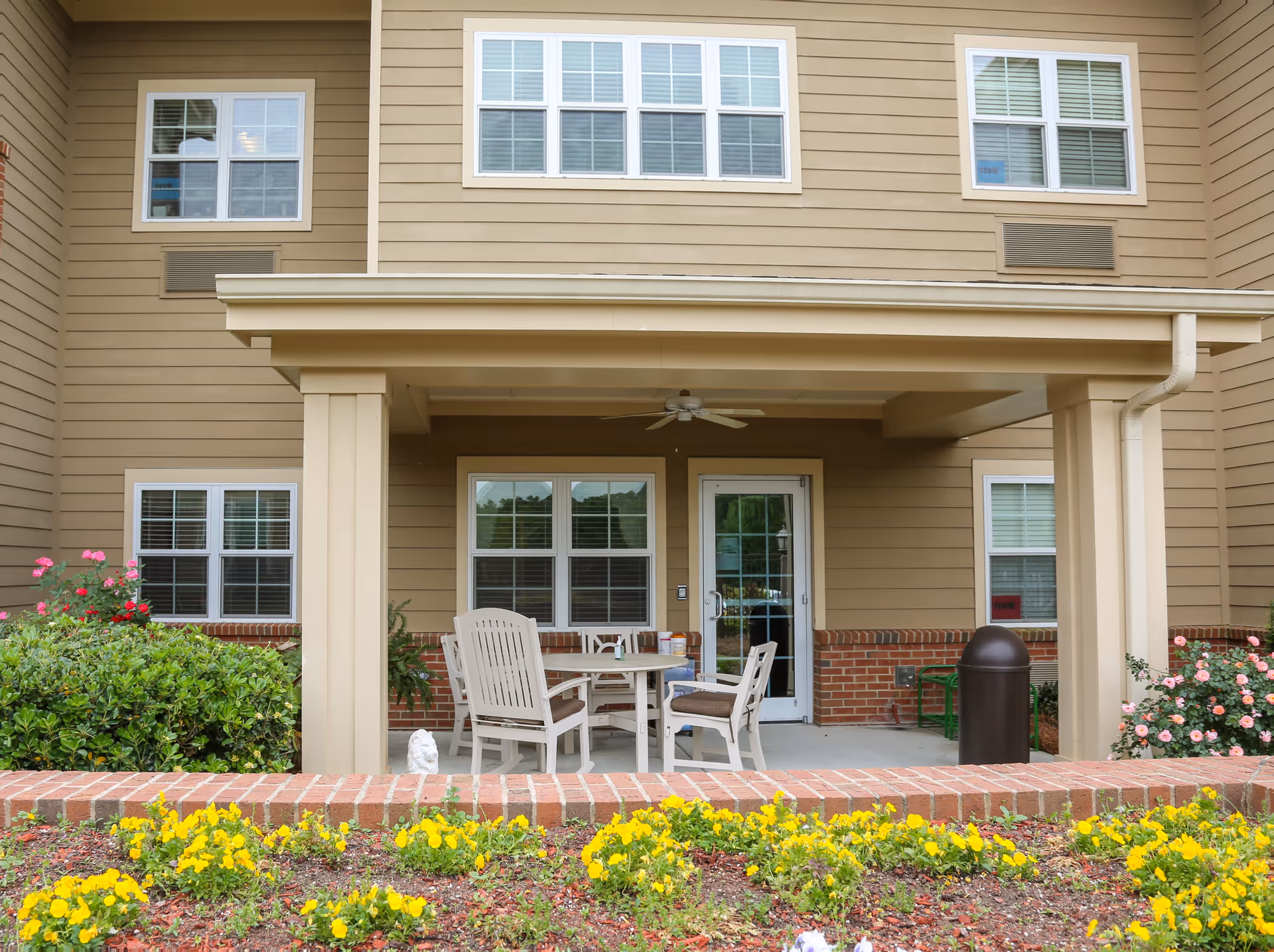 Covered outdoor patio area with a table and four chairs, surrounded by a garden with yellow flowers and bushes, attached to a beige building with multiple windows.