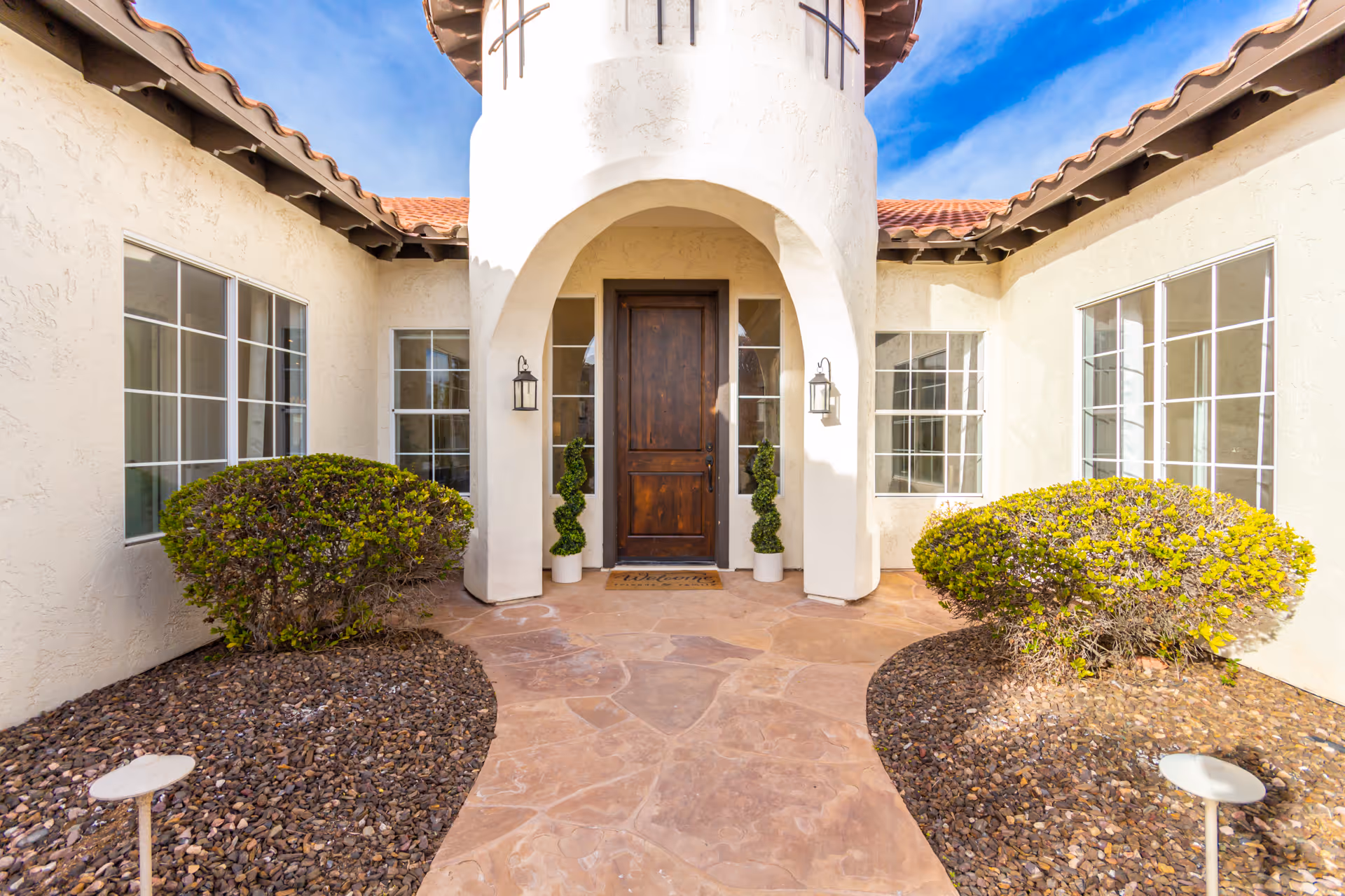 Entrance to a senior living facility with a wooden door set in a white stucco wall under an arched entryway. There are two potted topiary plants on either side of the door, flanked by two wall-mounted lantern-style lights. The pathway leading to the door is paved with stone tiles, bordered by small bushes and rock landscaping. The building has a tiled roof and multiple windows.