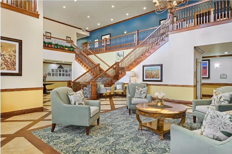 A spacious and well-lit common area with light blue armchairs arranged around a wooden coffee table on a patterned rug. The room features a large wooden staircase with railings leading to an upper floor, framed artwork on the walls, and a mix of cream and yellow paint with wood trim. The area has a welcoming and comfortable atmosphere.