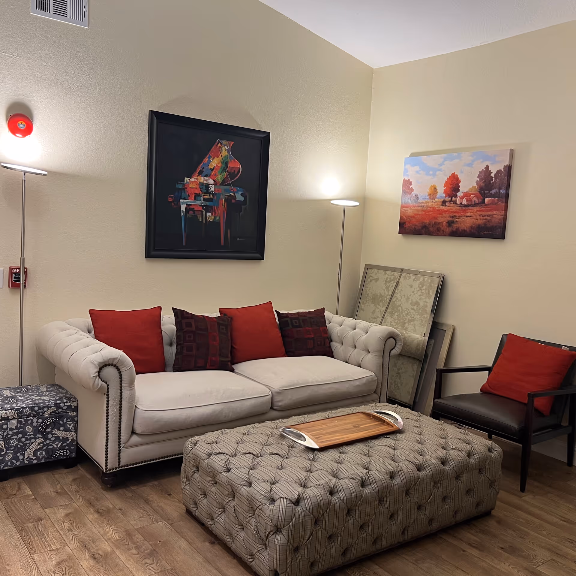 A cozy living room with a beige tufted sofa adorned with red and patterned cushions, a large tufted ottoman with a wooden tray on top, a dark armchair with a red cushion, two floor lamps, and two paintings on the walls. The floor is wooden, and there are some framed pictures leaning against the wall.