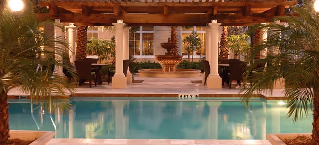 A serene outdoor courtyard area featuring a swimming pool with a depth marker of 4 feet 3 inches. The pool is surrounded by tropical plants and palm trees. In the background, there is a covered seating area with wooden beams and white columns, along with a decorative tiered fountain and additional greenery.