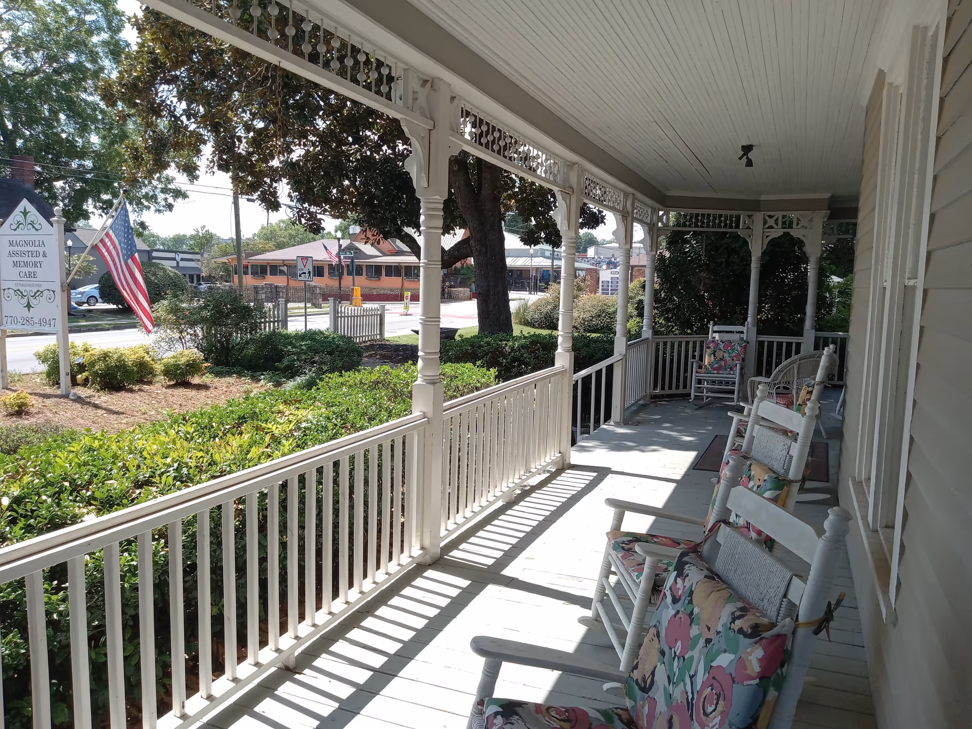 A sunny porch at Magnolia House with white wooden railings and columns, several white rocking chairs with floral cushions, and a view of a garden with bushes and trees. An American flag and a sign for Magnolia Assisted & Memory Care are visible near the street.