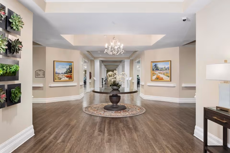 A spacious, well-lit hallway in a senior living facility with wooden flooring and beige walls. A round wooden table with a floral arrangement sits on a decorative rug in the center. The hallway is adorned with framed landscape paintings on the walls and a chandelier hanging from the ceiling. There are multiple doorways and a security camera visible in the upper right corner.