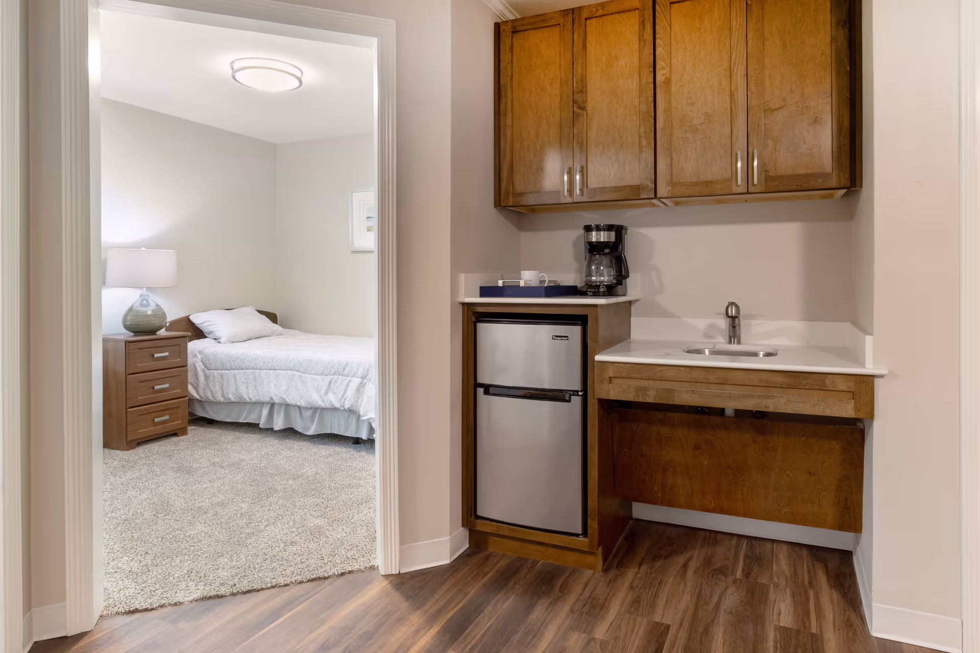 Small kitchenette with wooden cabinets, mini fridge, sink and coffee maker next to a doorway showing a bedroom with a bed and nightstand.