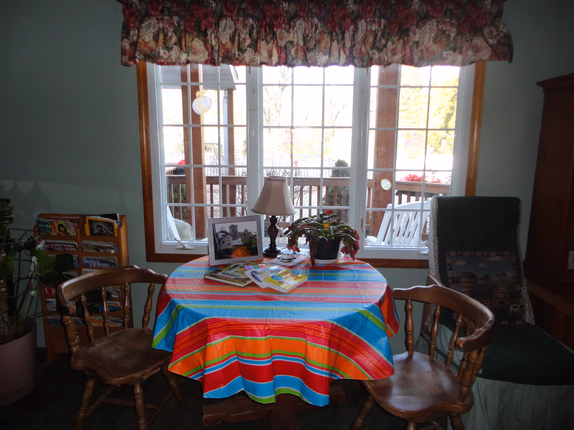 A cozy interior room with a round table covered by a colorful striped tablecloth. On the table are a small lamp, a potted plant, and some magazines or books. Two wooden chairs are positioned around the table. Behind the table is a large window with a floral valance, showing an outdoor porch with white rocking chairs and wooden railings.