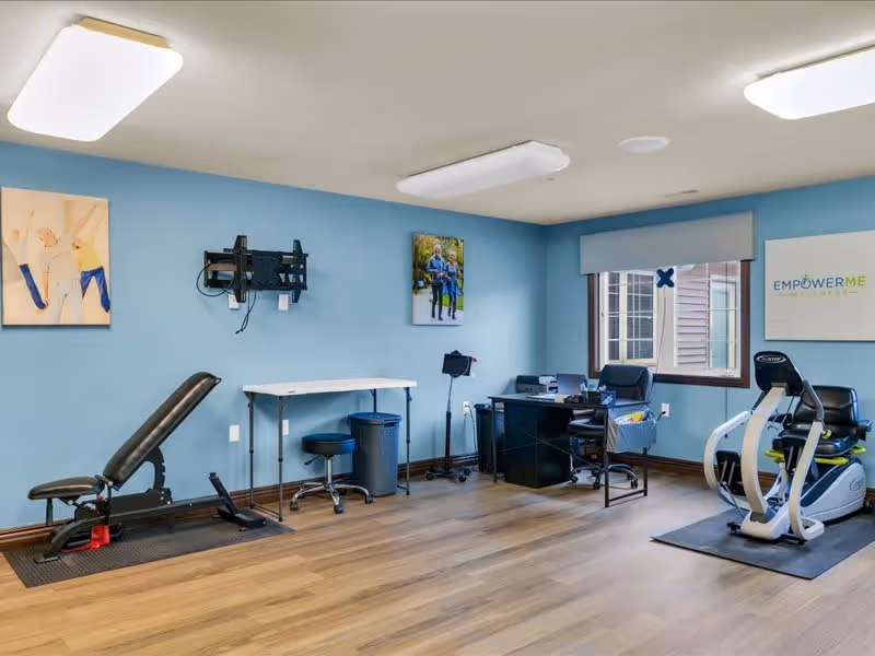 A wellness room with light blue walls and wood flooring featuring exercise equipment including a black adjustable bench and a recumbent exercise bike. There is a black desk with office supplies, a small white table with a stool, and a window with a view outside. The walls have framed pictures and a sign that reads 'EMPOWER ME WELLNESS'.