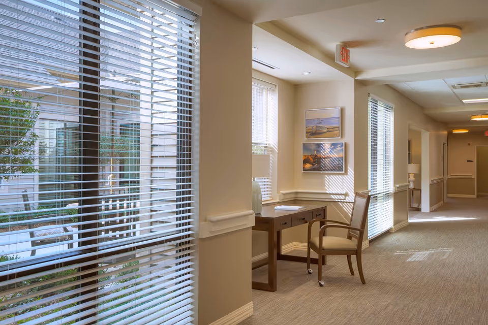 A well-lit hallway in a senior living facility with large windows covered by white blinds, a wooden desk with a chair, decorative paintings on the wall, and soft beige carpeting. The hallway extends into the distance with additional lighting fixtures on the ceiling.