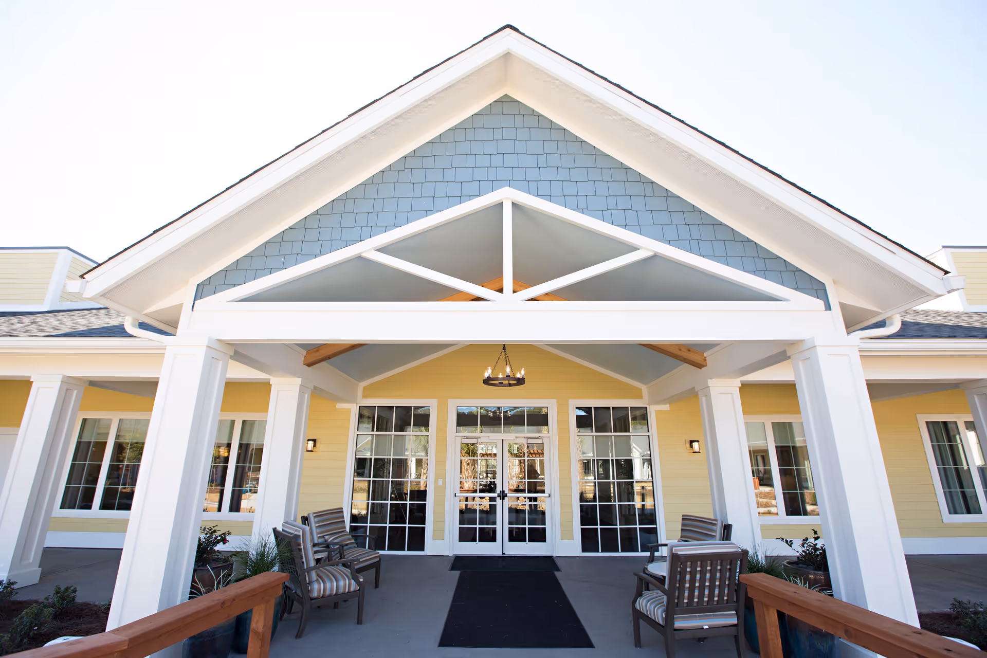 Front entrance of a senior living facility with a covered porch supported by white columns, yellow exterior walls, large windows, and a glass double door. There are several cushioned chairs arranged on the porch under a chandelier.
