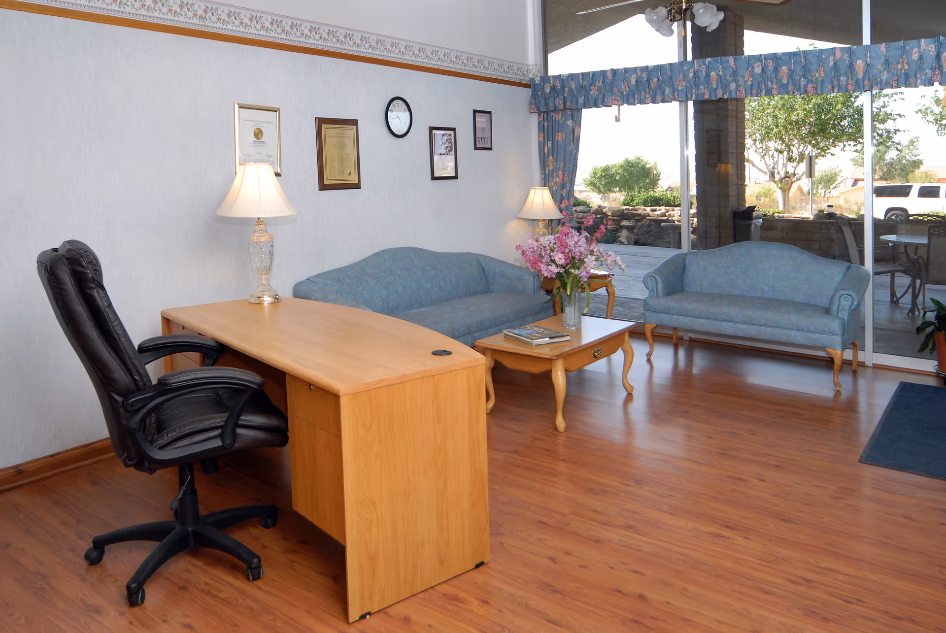 A waiting area with two blue upholstered sofas, a wooden coffee table with a flower vase and magazines, a wooden desk with a black office chair, two table lamps, framed certificates and a clock on the wall, and large glass windows showing an outdoor patio with trees and parked vehicles.