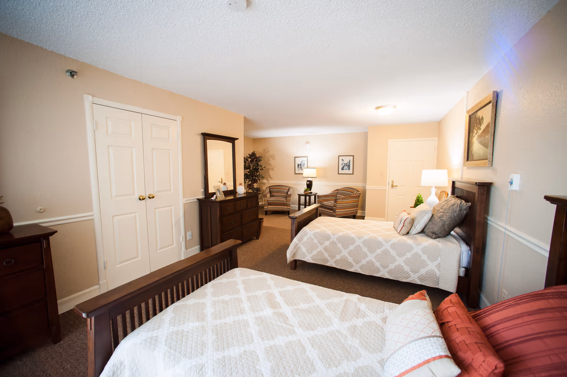 A senior living facility bedroom with two beds featuring patterned bedspreads and multiple pillows. The room includes wooden dressers, a mirror, two armchairs with a small table and lamp between them, framed pictures on the walls, and a door in the background. The walls are painted beige with white trim, and the room is well-lit with ceiling and table lamps.