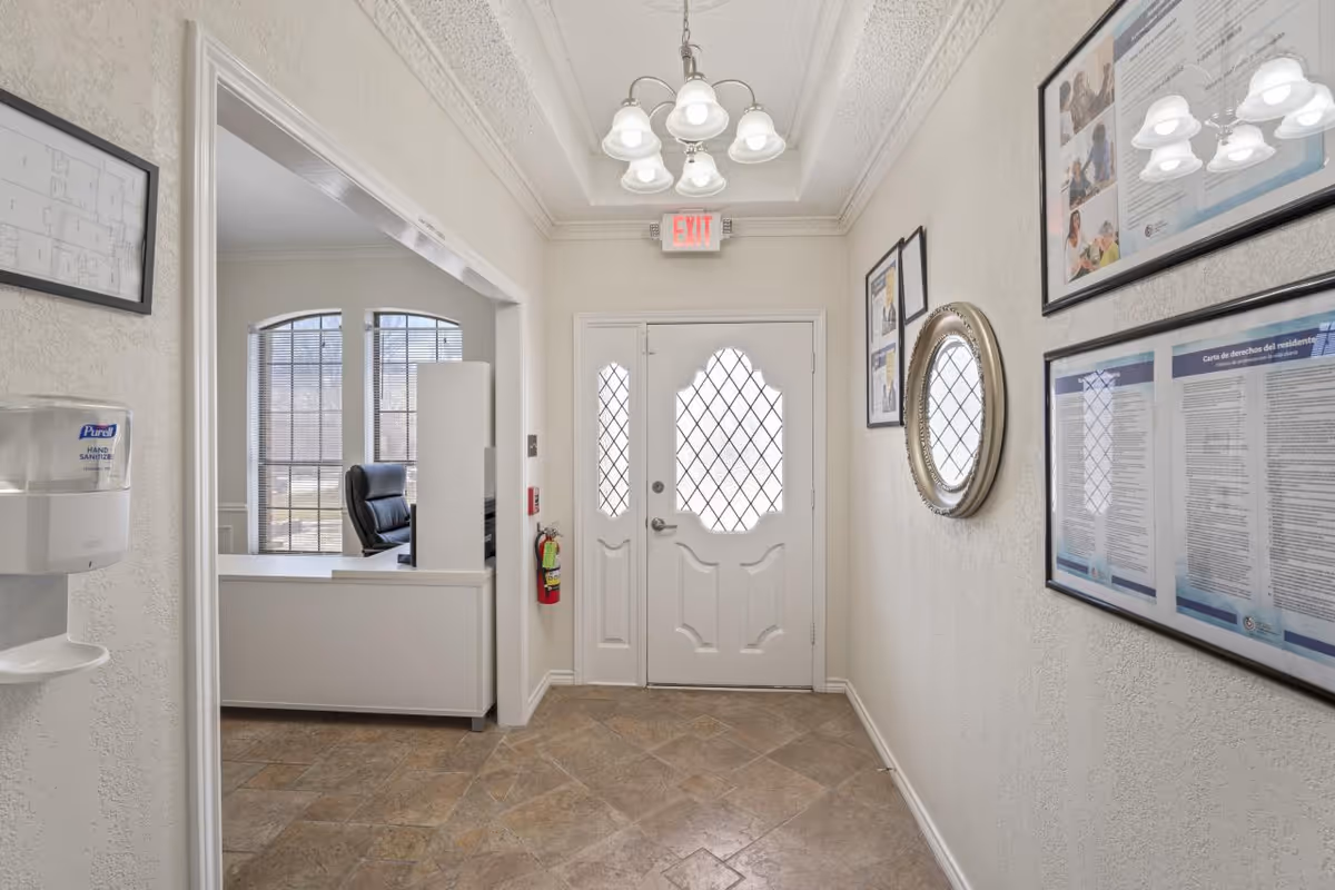 Interior view of an assisted living facility entrance area with a white door featuring decorative glass panels. To the left is a reception desk with a black office chair and large windows with blinds. The walls are light-colored with framed documents and a round mirror. A chandelier with multiple lights hangs from the ceiling, and a hand sanitizer dispenser is mounted on the left wall near the entrance to the reception area.