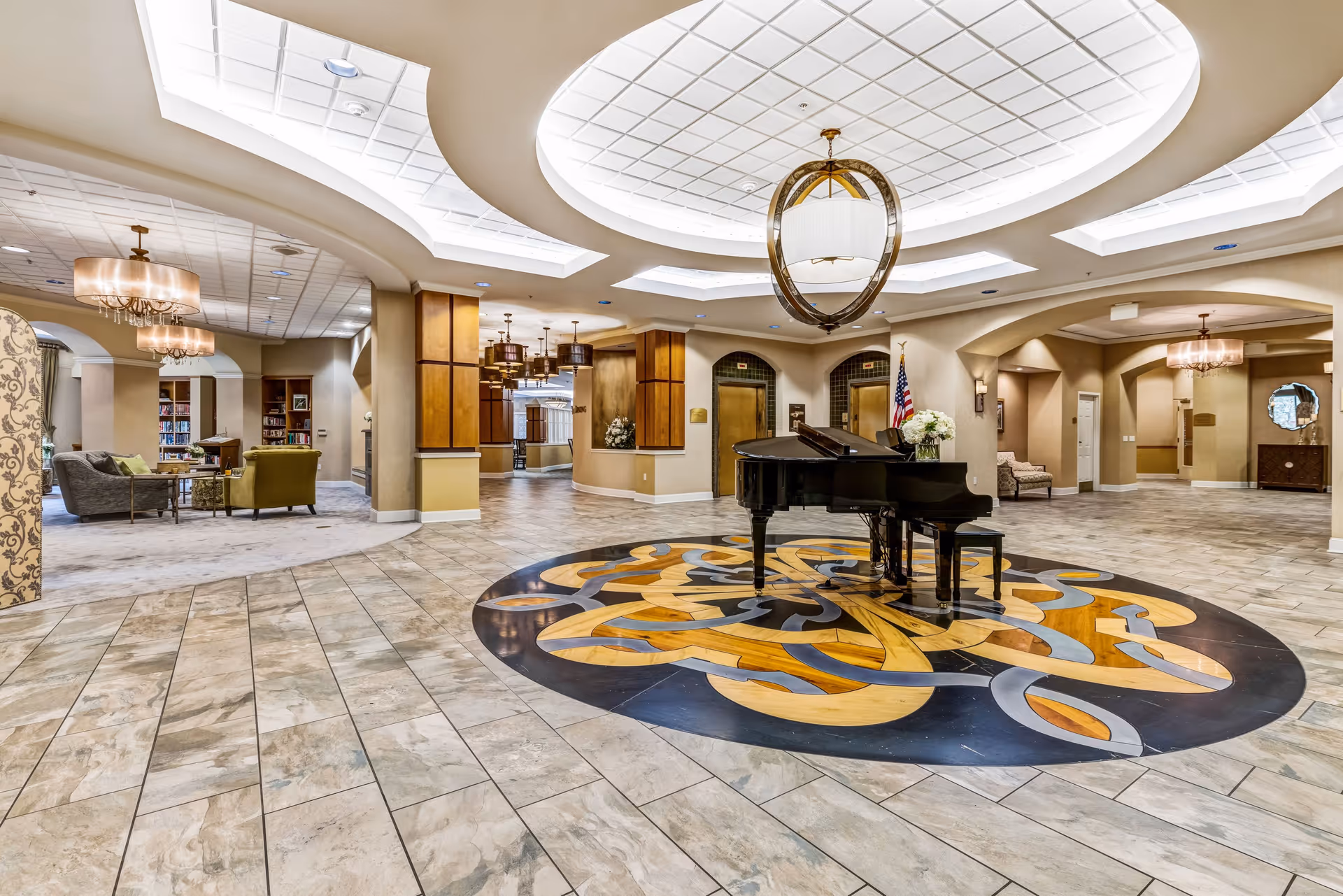 Spacious senior living facility lobby with a grand piano placed on an artistic circular floor design. The area features tiled flooring, elegant ceiling lights, and comfortable seating areas with sofas and armchairs. The space is well-lit with a combination of natural and artificial lighting, and there are decorative elements including flowers and an American flag near the elevators.