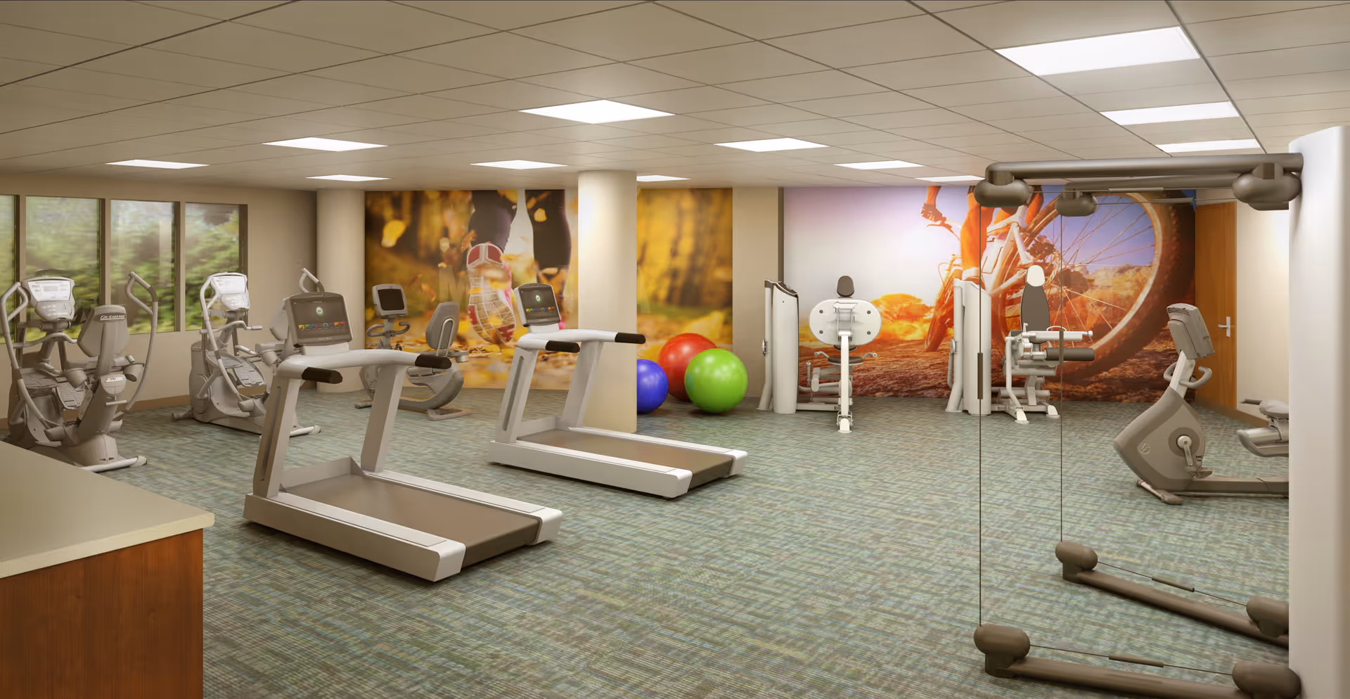 A spacious fitness room with exercise equipment including treadmills, stationary bikes, and weight machines. The room has large windows on one side, colorful exercise balls, and motivational wall murals depicting outdoor activities like running and biking. The floor is carpeted, and there is a mirrored wall on the right side.