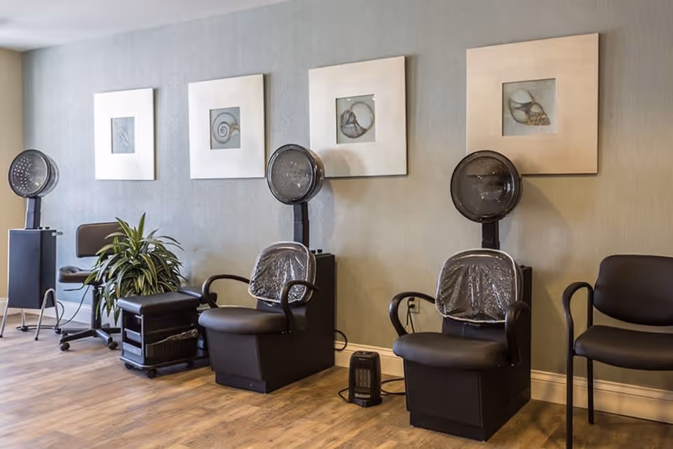 Interior of a senior living facility hair salon area with three black salon chairs equipped with hair dryers, a small black rolling cart, a potted plant, and four framed pictures of seashells on the wall.