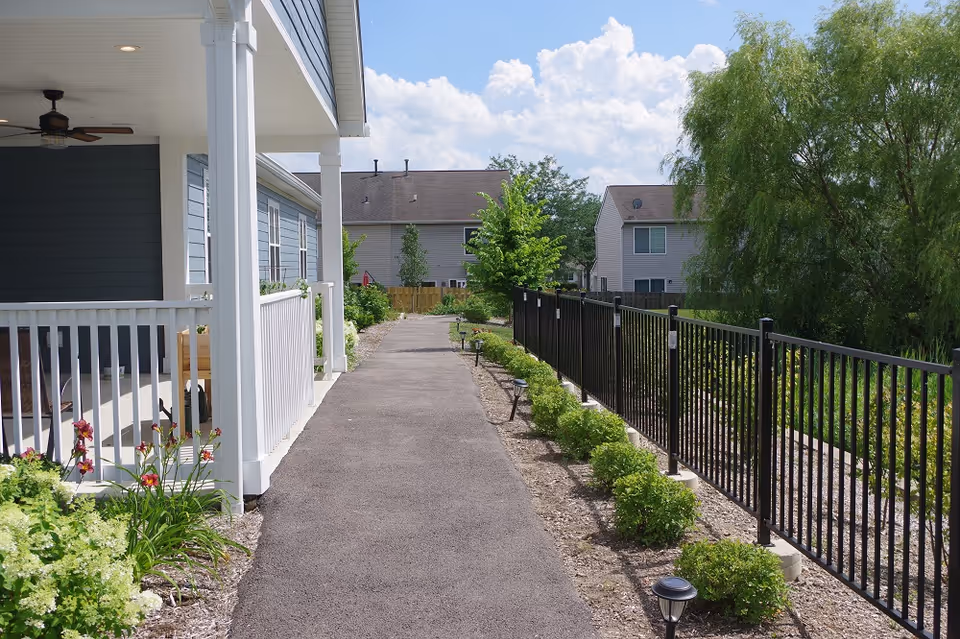 A paved walkway beside a building with a white porch railing and ceiling fan. On the right side, there is a black metal fence with small bushes and garden lights along the path. In the background, there are residential houses and trees under a partly cloudy sky.