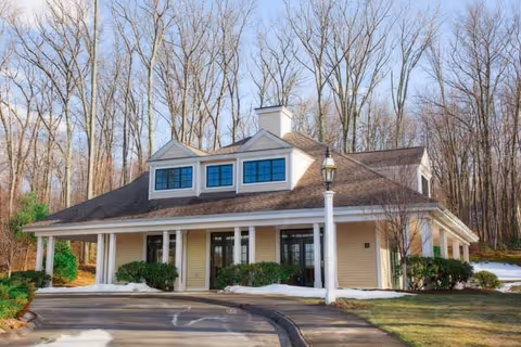 Single-story beige community building with a covered columned entrance, dormer windows, and a circular driveway framed by leafless trees.