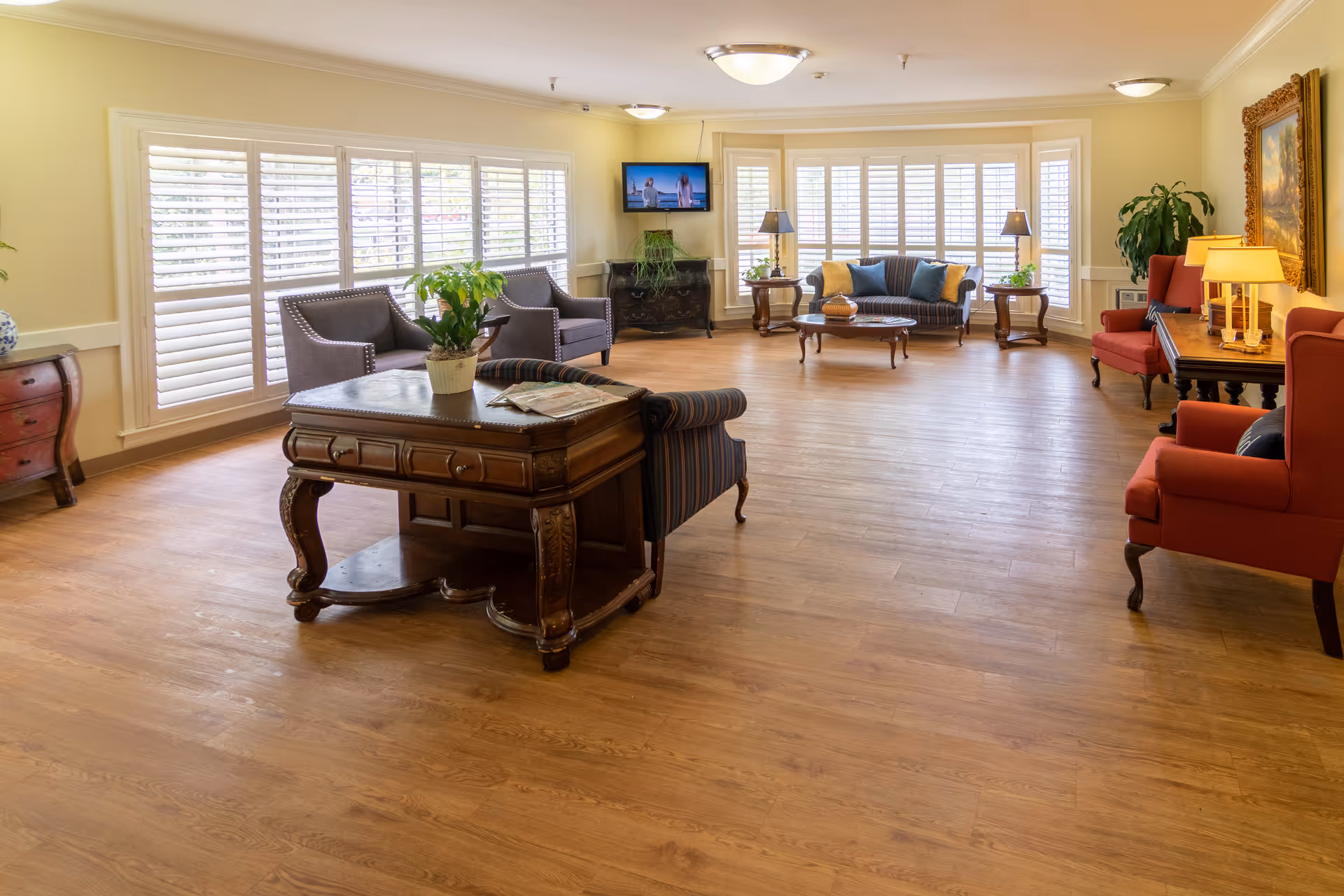 A spacious and well-lit living room area with wooden flooring, large windows with white shutters, multiple seating options including armchairs and sofas, a wooden coffee table, side tables with lamps, a wall-mounted TV, and decorative plants and artwork.