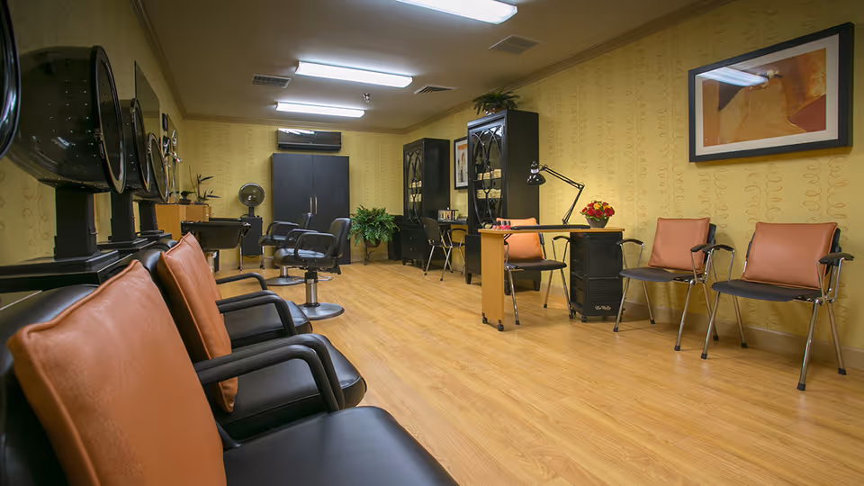Interior view of a senior community salon with multiple black salon chairs, hair dryers mounted on stands, a manicure table with chairs, and yellow patterned wallpaper. The room has wooden flooring, black cabinets, plants, and framed artwork on the walls.