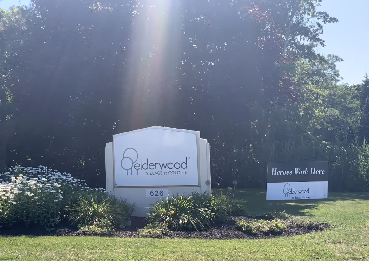 Entrance sign reading "Elderwood Village at Colonie" on a landscaped lawn with a smaller "Heroes Work Here" sign nearby.