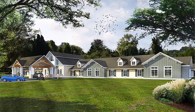 Single-story senior living building with pitched roofs and a covered entrance on a large green lawn surrounded by trees.