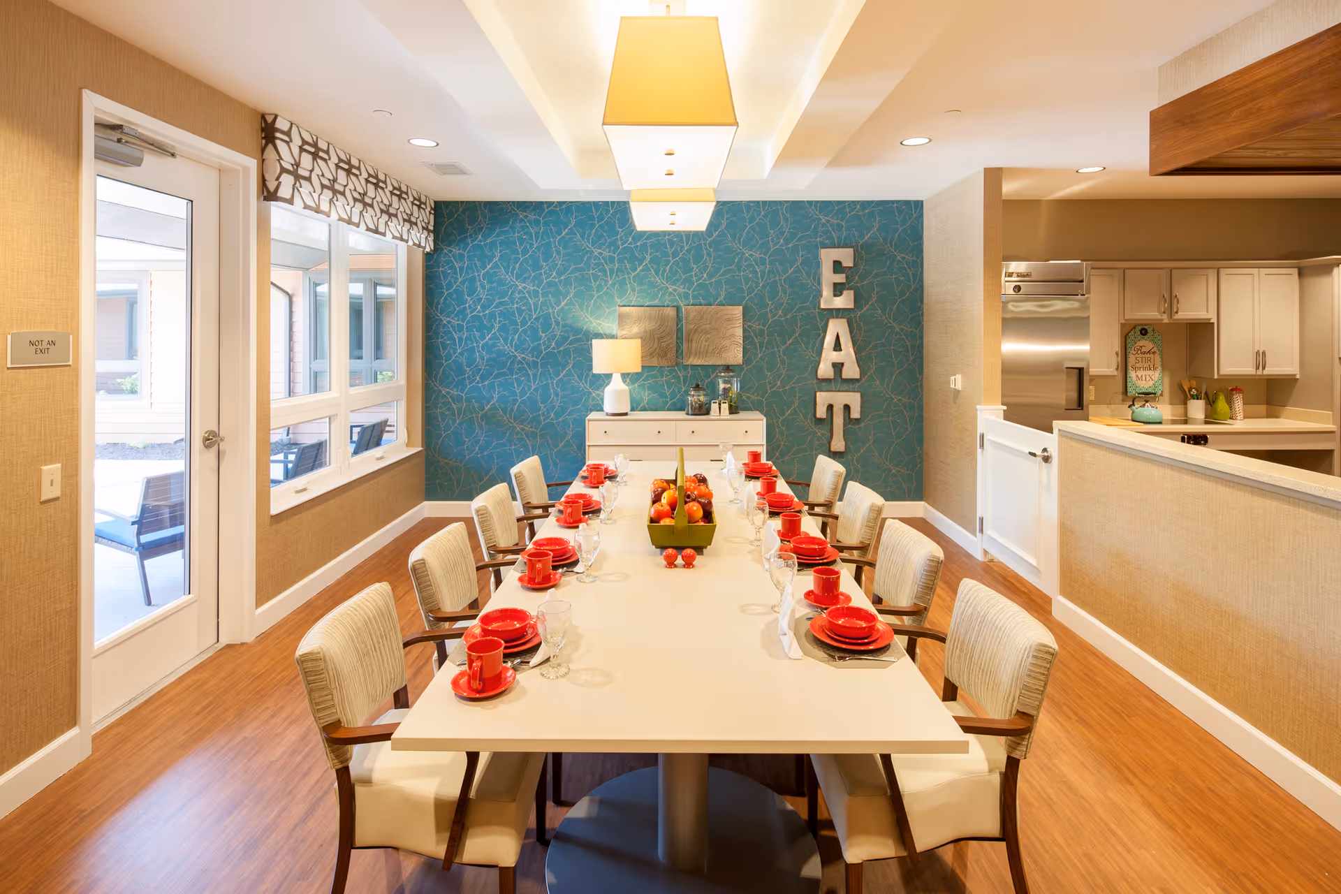 A bright dining room with a long white table set with red plates, cups, and glassware. The table is surrounded by beige cushioned chairs with wooden arms. On the far wall, there is a blue accent wall with a decorative pattern, a white sideboard with a lamp and decor items, and large letters spelling 'EAT'. To the right, there is a partial view of a kitchen area with white cabinets and stainless steel appliances. To the left, there is a door and windows looking out to an outdoor patio area with chairs.
