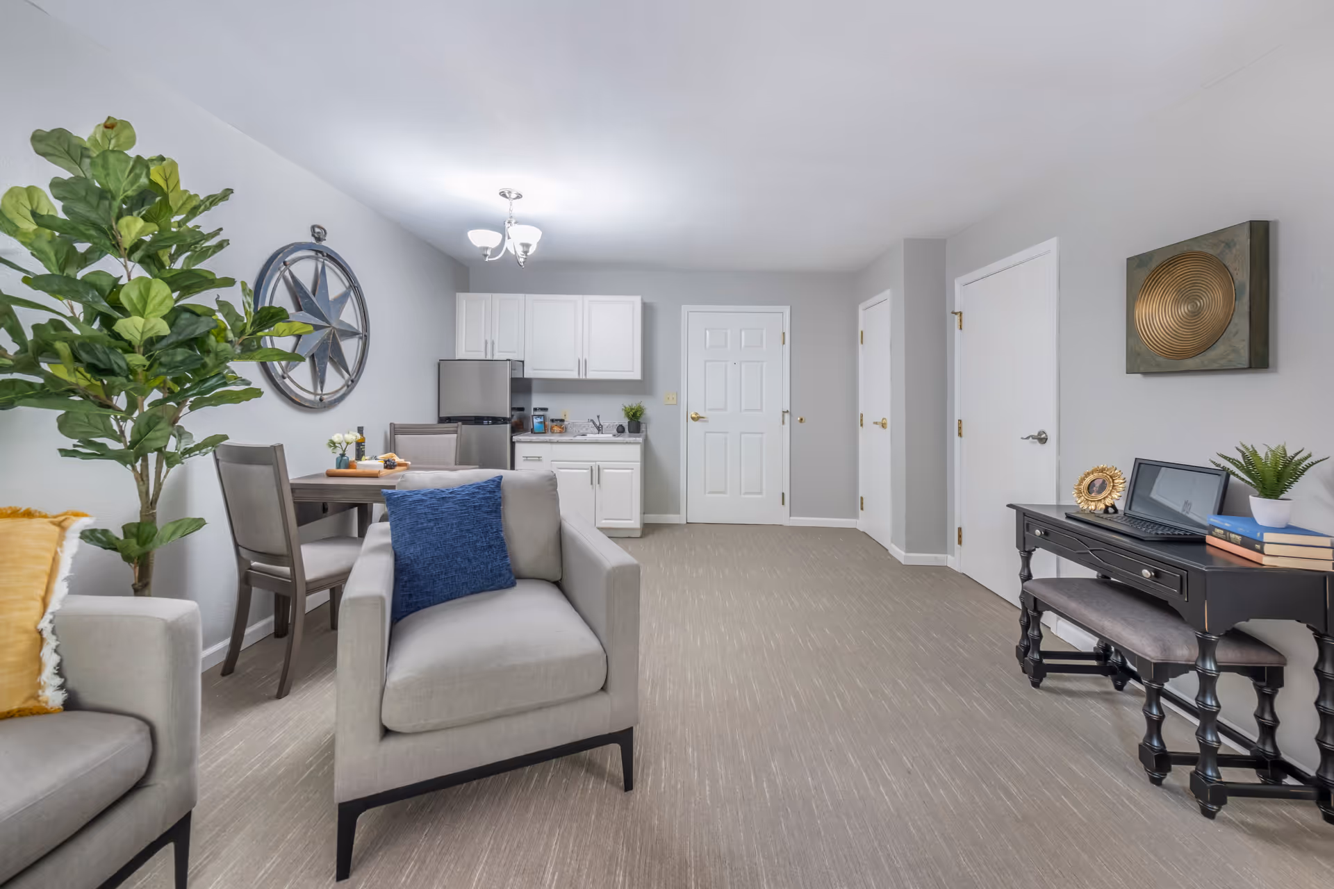 A bright and modern living area featuring a light gray armchair with a blue cushion, a small dining table with two chairs, a kitchenette with white cabinets and a mini refrigerator, a black desk with a laptop, books, and a small plant, and decorative wall art including a large compass and a circular gold piece. The room has light gray walls and carpeted flooring.