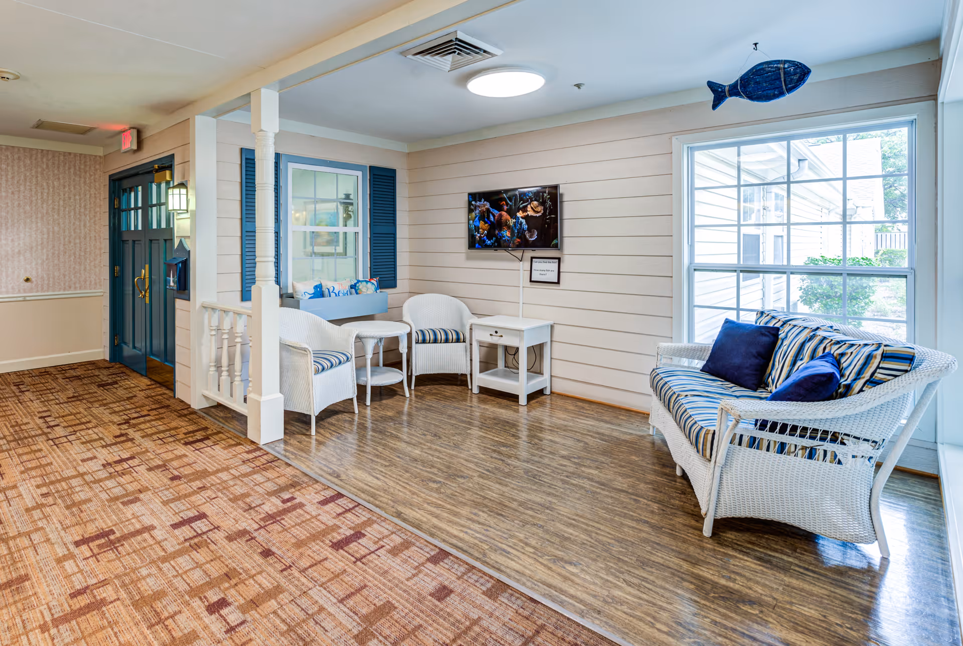 A cozy seating area in a senior living facility with white wicker furniture including a loveseat and two chairs with blue and white striped cushions. A small white table holds a flat-screen TV mounted on the wall displaying an underwater scene. The walls are light beige with blue window shutters and a large window letting in natural light. The floor transitions from patterned carpet to wood-like flooring in the seating area. A blue fish decoration hangs from the ceiling.