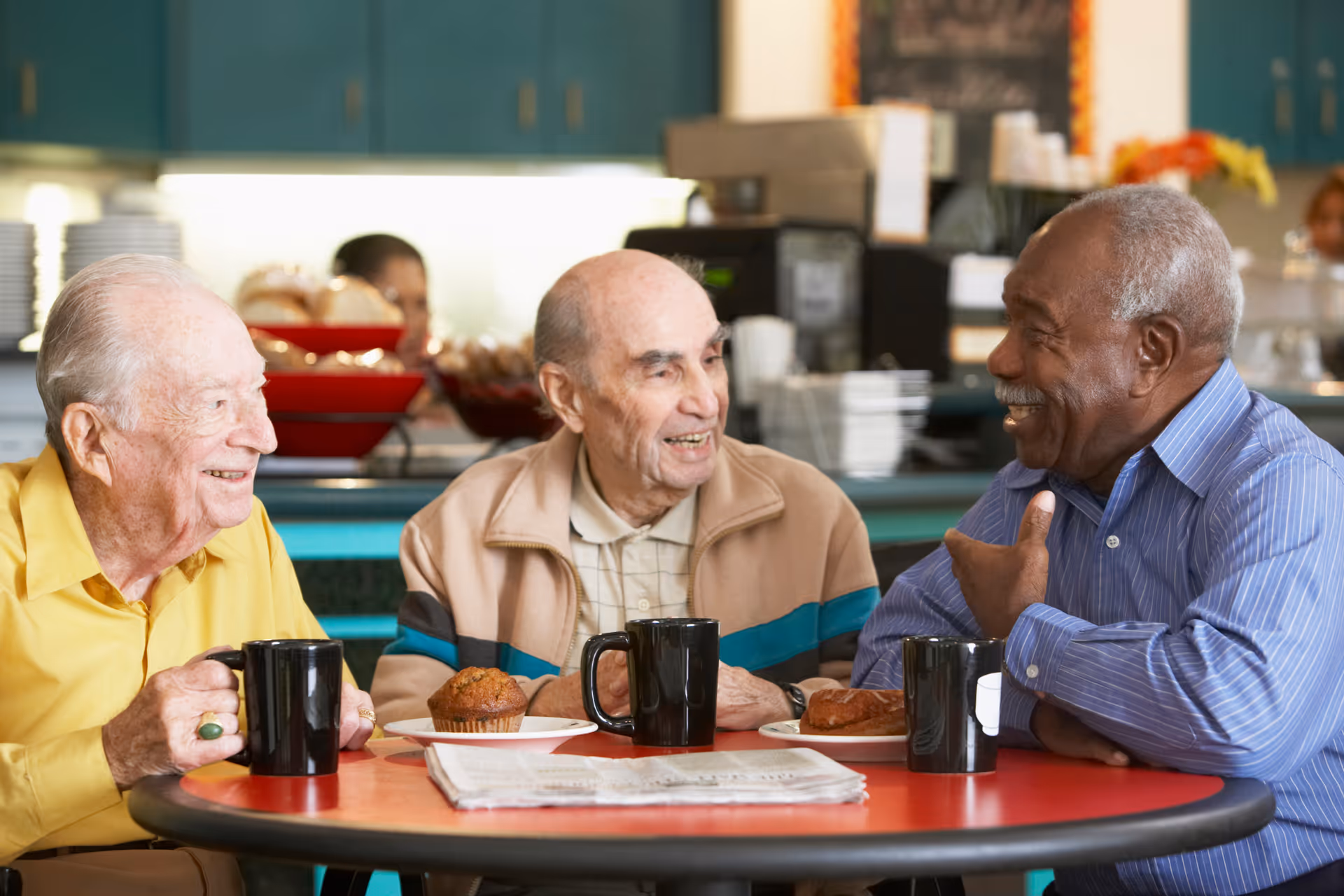 Three elderly men sitting around a round table in a dining area, smiling and chatting. Each has a black coffee mug and a plate with a muffin or pastry in front of them. The background shows a kitchen or cafeteria setting with blue cabinets and food items.