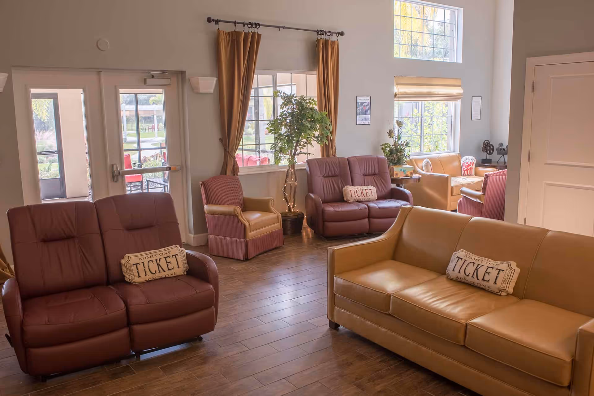 A bright and spacious living room area with multiple leather sofas and armchairs arranged around the room. The sofas have decorative pillows shaped like tickets. Large windows with curtains and blinds allow natural light to fill the space. There are plants near the windows and a door leading outside.